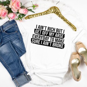 May include: White t-shirt with the text "I ain't rich but I get off my arse everyday to make sure I ain't broke" in black. The shirt is displayed with blue jeans, a gold hanger, and high heels.