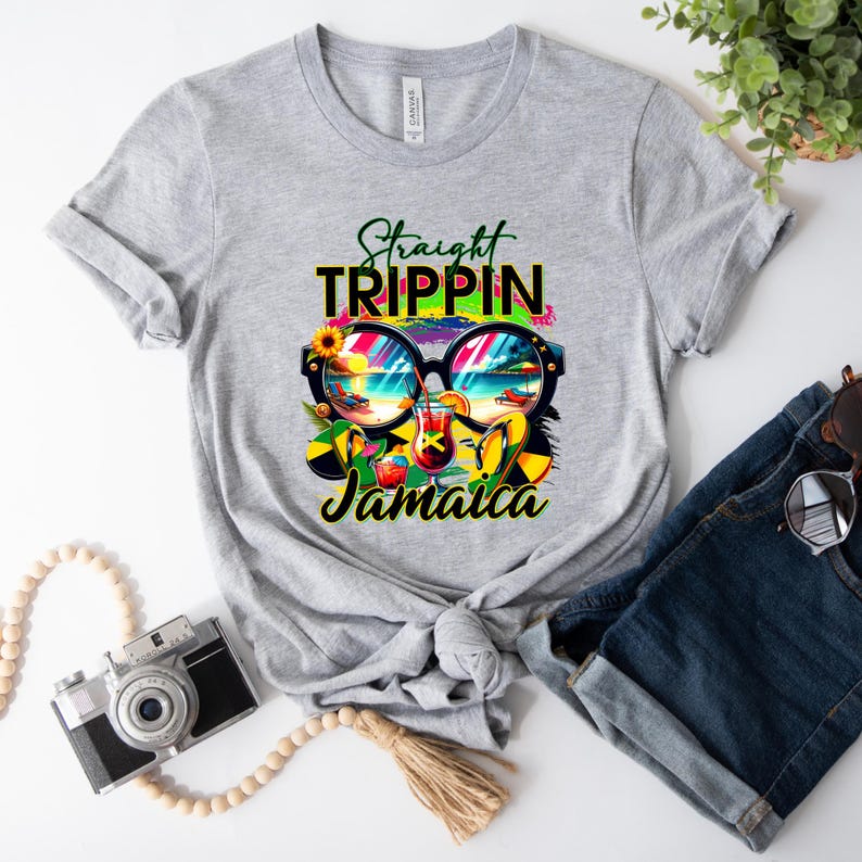 May include: A light gray t-shirt featuring the words "Straight Trippin' Jamaica" in a colorful design. The graphic includes sunglasses reflecting a beach scene, cocktails, and Jamaican flag elements. The shirt is paired with denim shorts.