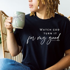 Puede incluir: Camiseta negra con el texto "Watch God Turn It for my good" en escritura blanca. Una taza verde claro con un diseño de libélula se sostiene en la mano de la persona.