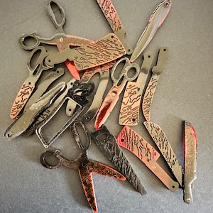 May include: A collection of black and red patterned knife and scissor blades. The blades have a decorative pattern and are arranged in a pile.