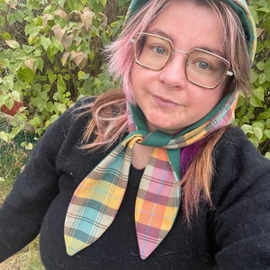 Peut inclure: Un ensemble écharpe et capuche à carreaux colorés. L'écharpe présente un motif vibrant de carrés jaunes, bleus, roses et oranges. La capuche est verte avec un motif à carreaux assorti. La personne porte un pull noir et des lunettes.