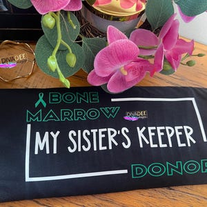 May include: Black t-shirt with white text that reads "Bone Marrow My Sister's Keeper Donor". The text is framed in a white rectangle. A green ribbon is above the text.