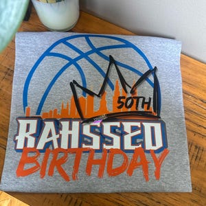 May include: A gray t-shirt with a basketball graphic in blue, a crown, and the text "50th". The shirt also features the name "RAHSSED" and the word "BIRTHDAY" in orange and white lettering. The design has a sporty theme.