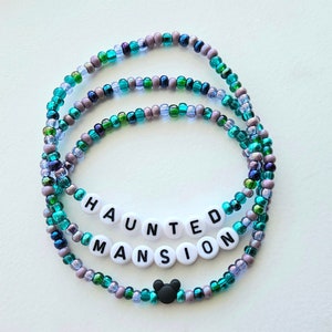 May include: A beaded bracelet featuring a mix of translucent teal, green, and metallic purple glass beads. The bracelet includes white circular beads with black lettering spelling out HAUNTED MANSION and a small black mouse-shaped bead.