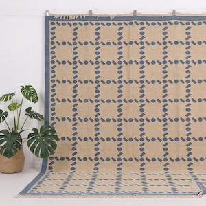 May include: A large beige rug with a geometric pattern of blue shapes. The rug has a blue border and is displayed against a white wall. A potted plant with large green leaves sits to the left of the rug.