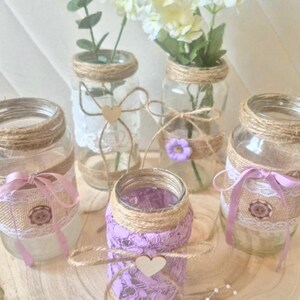 Peut inclure: Cinq bocaux en verre transparent décorés de ficelle, de dentelle et de rubans. Deux bocaux contiennent des fleurs blanches et de la verdure. Les bocaux présentent des détails comme un petit cœur en bois, une fleur violette et un emblème circulaire.