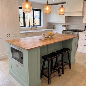 May include: A kitchen island with a light green base and a natural wood countertop. The island features a built-in microwave, two black wooden stools, and three ribbed glass pendant lights hanging above. The kitchen includes white cabinetry.