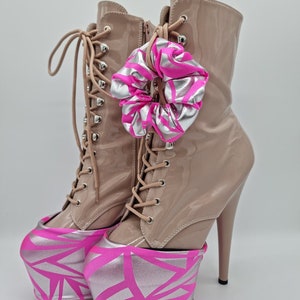 May include: A pair of beige platform boots with pink and silver geometric patterned fabric covers on the platform. The boots have lace-up fronts and a high heel. A pink and silver scrunchie is tied around the laces.