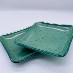 May include: Two square glass dishes with a smooth, glossy finish in a vibrant seafoam green colour. Each dish features a distinct speckled pattern with small, dark brown dots scattered across the surface, and slightly raised, rounded edges.
