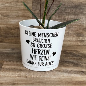Könnte beinhalten: Ein weißer Keramiktopf mit schwarzem Text, der "Kleine Menschen brauchen so grosse Herzen wie Deins! Danke fur alles" lautet. Der Text ist von zwei schwarzen Herzen umgeben. Eine kleine grüne Pflanze befindet sich im Topf.