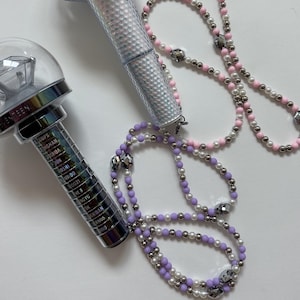 May include: Two light sticks with diamond-shaped tops and silver handles. One has a clear, faceted top and the other has a clear top with a diamond shape inside. Two beaded necklaces, one pink and white, the other purple and white, are also in the image.