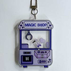 May include: A purple and white acrylic keychain shaped like a claw machine, with the words "MAGIC SHOP" at the top. Inside are small graphics of a microphone, heart, and other items. Includes a silver clasp.