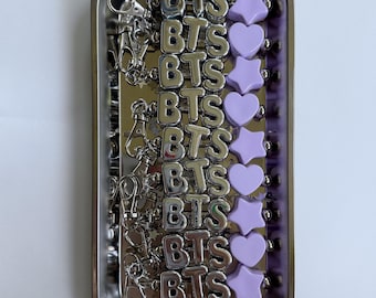 May include: A collection of silver-colored BTS charms and purple heart and star-shaped beads, displayed in a rectangular metal container. The charms are arranged in rows, with a clasp attached for easy use.