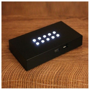 Op de afbeelding: Een zwarte rechthoekige displaystandaard met negen helderwitte LED-lampjes in een raster. Het heeft een USB-poort en een geheugenkaartsleuf. De standaard staat op een houten oppervlak.