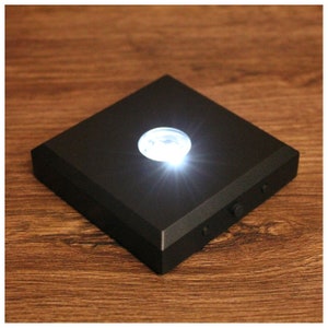 Può includere: Una base di luce LED quadrata nera con una piattaforma circolare trasparente in cima. La luce è accesa e illumina la piattaforma.