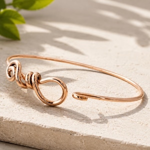 Op de afbeelding: Een roségoudkleurige metalen armband met een dunne, gepolijste draadband. Het ontwerp heeft aan één kant een decoratief middelpunt, bestaande uit een reeks strakke, cirkelvormige draadlussen en een grotere, druppelvormige draadlus.