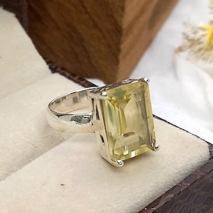May include: A silver ring featuring a large, pale yellow, emerald-cut gemstone. The ring is presented in a brown velvet ring box.