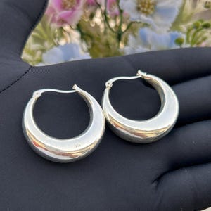 May include: A pair of polished silver hoop earrings with a thick, rounded, crescent-shaped design. The earrings feature a smooth, reflective metallic surface and a standard hinged post closure mechanism, showcasing a simple and classic circular form.