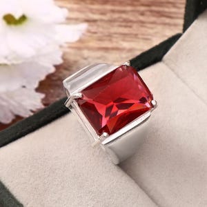 May include: A silver ring featuring a large, rectangular, faceted red gemstone. The ring is set in a simple, modern style, and is displayed in a black jewelry box with a white interior. White flowers are in the background.