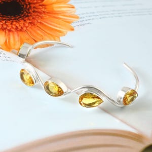 Op de afbeelding: Een zilveren armband met een golfontwerp, met vijf gele, druppelvormige edelstenen. De armband staat tegen een lichte achtergrond met een oranje bloem in de linkerbovenhoek.