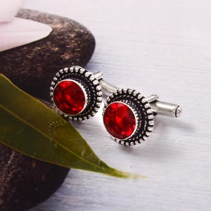May include: A pair of silver cufflinks with red gemstone accents. The cufflinks have a circular design with a textured border.