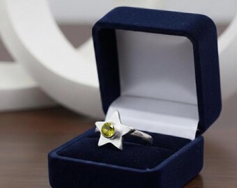 May include: A silver star-shaped ring with a round, light green gemstone, presented in a dark blue velvet ring box. The ring's star design is the focal point, with the gemstone adding a pop of color.