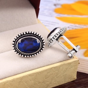 May include: A pair of silver cufflinks with oval blue gemstone accents. The cufflinks have a detailed silver design around the edges of the gemstones.