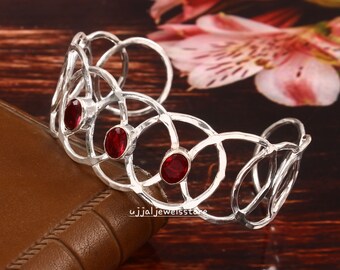 May include: Silver bangle bracelet with a delicate, openwork design featuring three red gemstones. The bracelet is made of hammered silver and has a slightly textured finish.