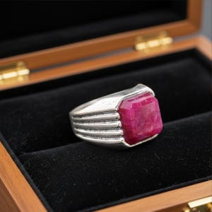 May include: A silver ring featuring a large, rectangular, bright red gemstone. The ring has a textured band with horizontal lines. The ring is set against a black velvet background inside a wooden box.