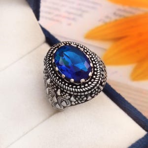 Peut inclure: Une bague en argent avec une pierre précieuse bleue de forme ovale sertie dans une bande en argent détaillée.