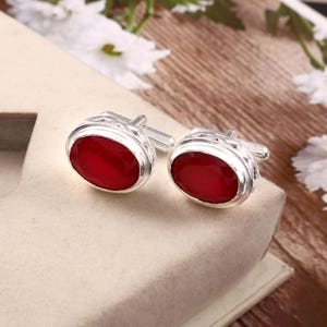 May include: Silver cufflinks featuring oval, faceted, red gemstones. The cufflinks are presented on a neutral surface, with a blurred background of white flowers and wood.