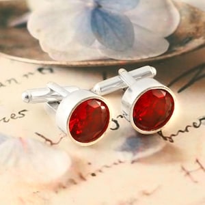 Peut inclure: Une paire de boutons de manchette argentés avec une pierre précieuse rouge vif et ronde sertie dans un cadre argenté. Les boutons de manchette sont présentés sur une surface à motifs avec un élément floral flou en arrière-plan. Un accessoire classique.