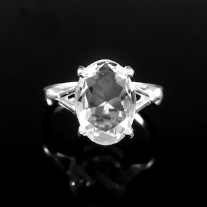 May include: A silver ring featuring a large, oval-cut, clear gemstone. The gemstone is the focal point, held in place by a four-prong setting. The ring band has a delicate, open design. The ring is set against a black background.