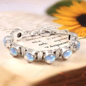 Peut inclure: Un bracelet en argent avec une fermeture à fermoir présente une rangée de perles de pierre de lune irisées. Le bracelet est présenté sur un livre avec un tournesol en arrière-plan.