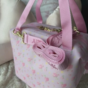 May include: A pink floral print handbag with a matching shoulder strap and gold-colored hardware. The bag features a rectangular shape with a top handle and a detachable strap. The floral pattern includes pink roses and yellow accents.