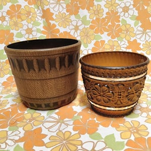 May include: Two brown ceramic plant pots. The larger pot has a textured, ridged design. The smaller pot features a floral pattern with a white band. Both pots are empty and sit on a floral patterned fabric.