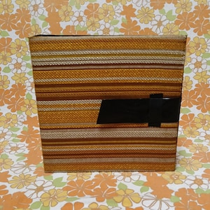 May include: A rectangular woven box with horizontal stripes in shades of brown, tan, and gold. A black rectangular accent is on the front. The box is set against a floral patterned background.