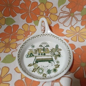 Może przedstawiać: Biała ceramiczna podstawka pod łyżkę z uchwytem, z zielono-brązową ilustracją osoby przy stole z jedzeniem i kwiatami. Obrzeże zdobione zielonym wzorem kwiatowym.