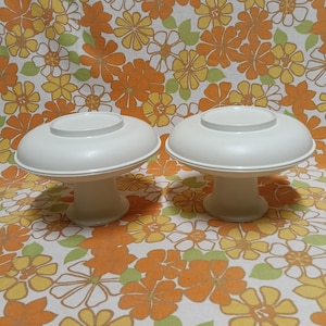 Könnte beinhalten: Zwei weiße Tortenständer mit Sockel, jeweils mit einer breiten, flachen Oberseite und einem abgerundeten Sockel. Sie stehen vor einem floral gemusterten Hintergrund in Orange, Gelb und Grün.