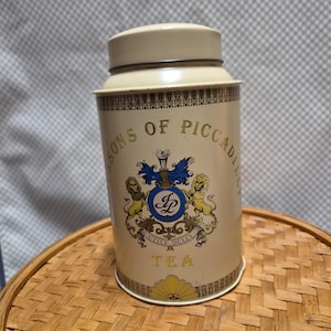 May include: A cylindrical cream-colored metal tea tin with a matching lid. The front features a crest with two lions, a blue circular emblem with the letters FP, and the text SONS OF PICCADILLY and TEA. A decorative yellow fan pattern borders the base.