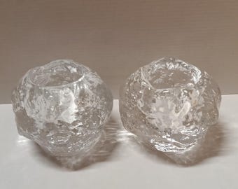 May include: Two clear, textured glass candle holders. Each holder has a rounded shape with a central opening for a candle. The glass has a rough, organic texture, giving them a unique, natural appearance.