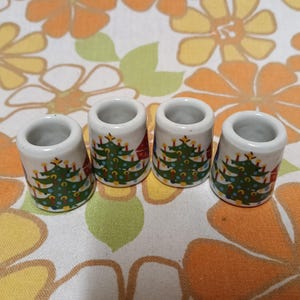 May include: Four white ceramic thimbles with a Christmas tree design. Each thimble features a green Christmas tree decorated with red and yellow ornaments, and a small red house. The thimbles are arranged on a floral patterned fabric.