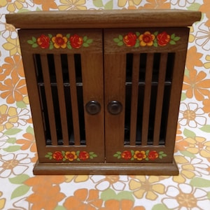 Peut inclure: Un petit cabinet en bois avec deux portes, peint en brun avec des détails floraux en rouge, jaune et vert. Le cabinet a un design à lattes sur les portes et deux boutons noirs.