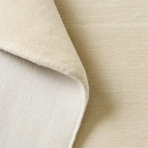 May include: A solid cream-colored area rug with a soft, dense pile texture. The rug is folded over at one corner, revealing the lighter-toned underside and the thick, plush construction of the fabric surface across the entire visible area.