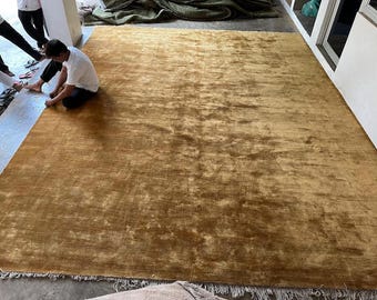 May include: A large, rectangular area rug in a solid mustard gold color with a soft, velvet-like texture. The rug features a short, dense pile and a fringe trim along the bottom edge, displaying subtle variations in light and shadow across its surface.