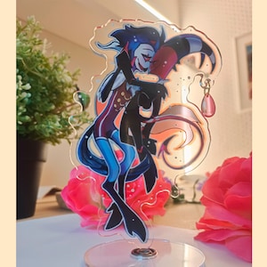 May include: A clear acrylic stand with a colorful illustration of two cartoon characters hugging. The characters are a blue and black demon with horns and a red demon with a long tail. The stand has a sparkly background and a pink dangle earring.