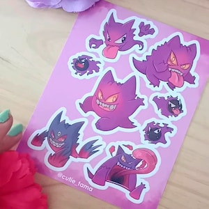 Puede incluir: Un conjunto de ocho pegatinas con diferentes ilustraciones del Pokémon Gengar. Las pegatinas están sobre un fondo rosa con un efecto acuarela. Las pegatinas son de diferentes tamaños y presentan a Gengar en diferentes poses. El texto "@cutie_tama" está en la parte inferior de la hoja.