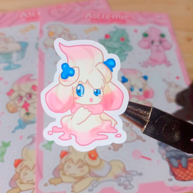 Puede incluir: Una pegatina troquelada con un personaje de dibujos animados con un cuerpo color crema, detalles en rosa y ojos azules. El personaje tiene una bufanda rosa y est&aacute; rodeado de una sustancia rosa similar al helado derretido. Otras pegatinas con dise&ntilde;os similares est&aacute;n en el fondo.
