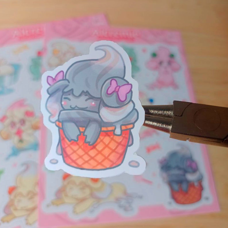 Puede incluir: Una pegatina con un personaje de cono de helado de dibujos animados con helado suave gris y blanco, lazos rosas y una cara sonriente. El cono es naranja con un patr&oacute;n de gofres. Otras pegatinas son visibles en el fondo.