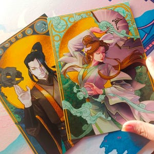 Puede incluir: Dos tarjetas ilustradas con un borde de estilo art déco. La tarjeta de la izquierda presenta a un hombre con cabello negro y un atuendo negro y dorado sosteniendo una calavera negra. La tarjeta de la derecha presenta a una mujer con cabello largo castaño que lleva un atuendo blanco y morado con un abanico en la mano. El texto en el abanico está en chino.